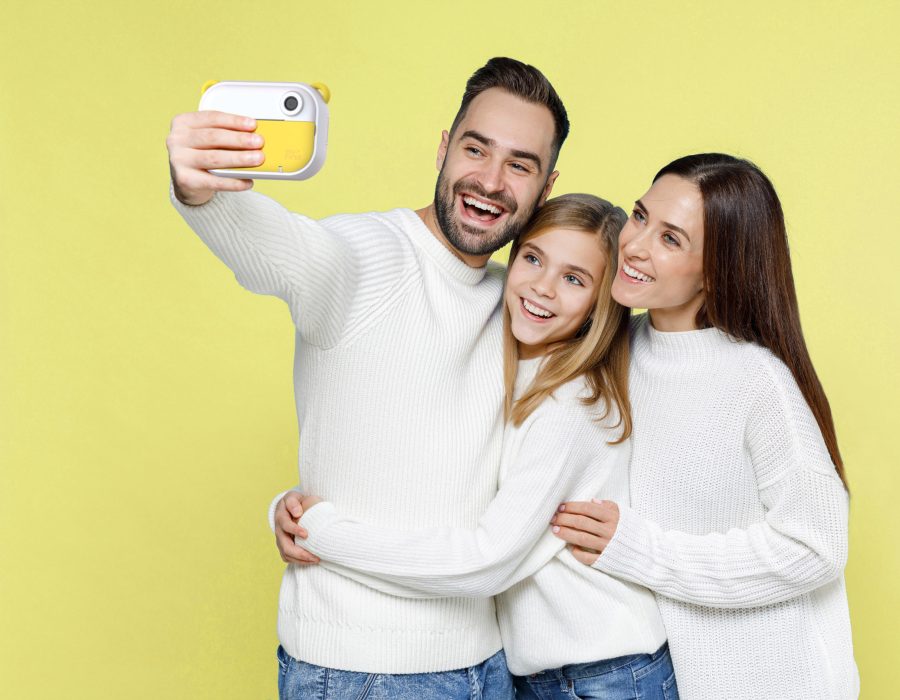 Happy young parents mom dad with child kid daughter teen girl in white sweaters doing selfie shot on mobile phone hugging isolated on blue background studio. Family day parenthood childhood concept